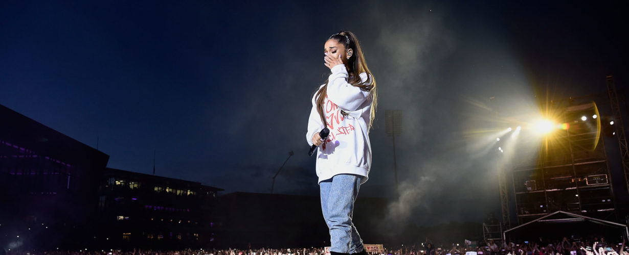 Musician Ariana Grande stands on stage in front of audience, wiping away a tear
