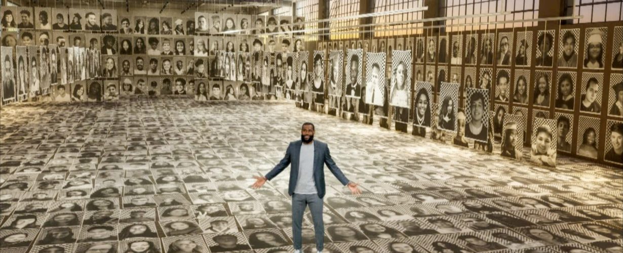 LeBron James stands with his hands open surrounded by sepia-toned portraits of graduating high school seniors